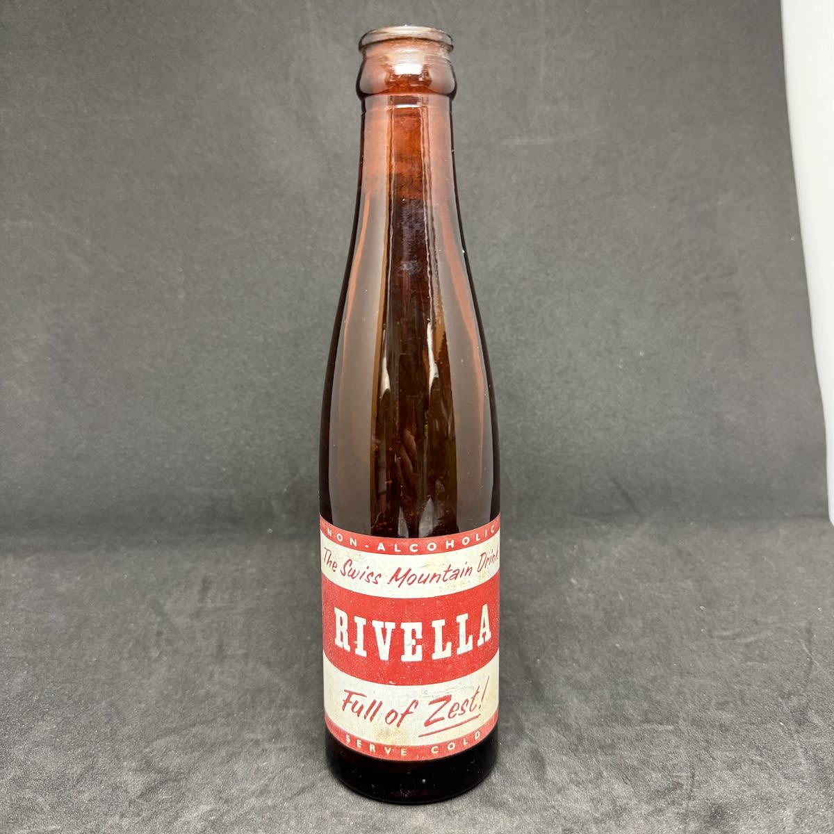 Ceramic Label Crown Seal. Rivella The Swiss Mountain Drink. Rivella (Australia) Pty Ltd. Amber. Red and white print. 8 oz.