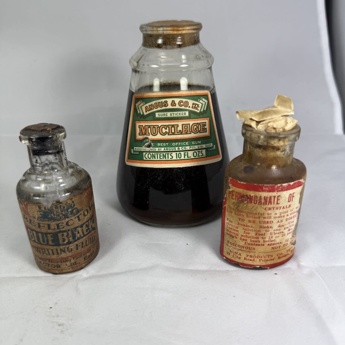 3 small labelled bottles: Angus & Co Mucilage (sealed with contents) Reflector Ink, Brisbane Permanganate of...Iona Products Melbourne.