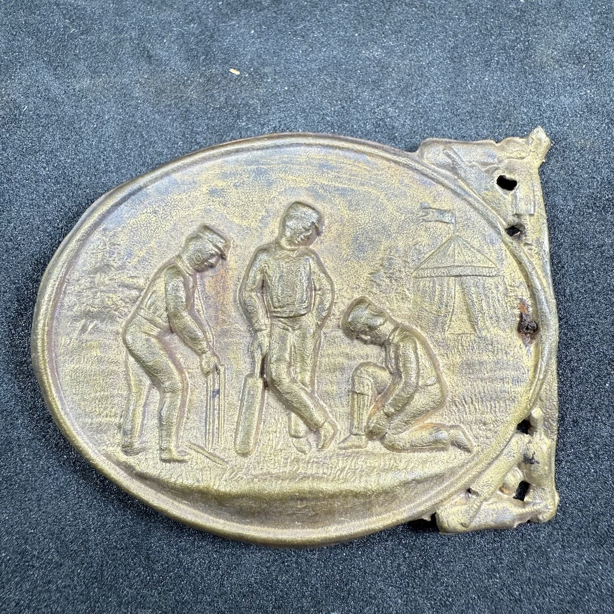 Cricket Belt Sash Buckle. Three players: One at the stumps, a batsman leaning on his bat and looks like the third player is putting on pads. Tent in Background.