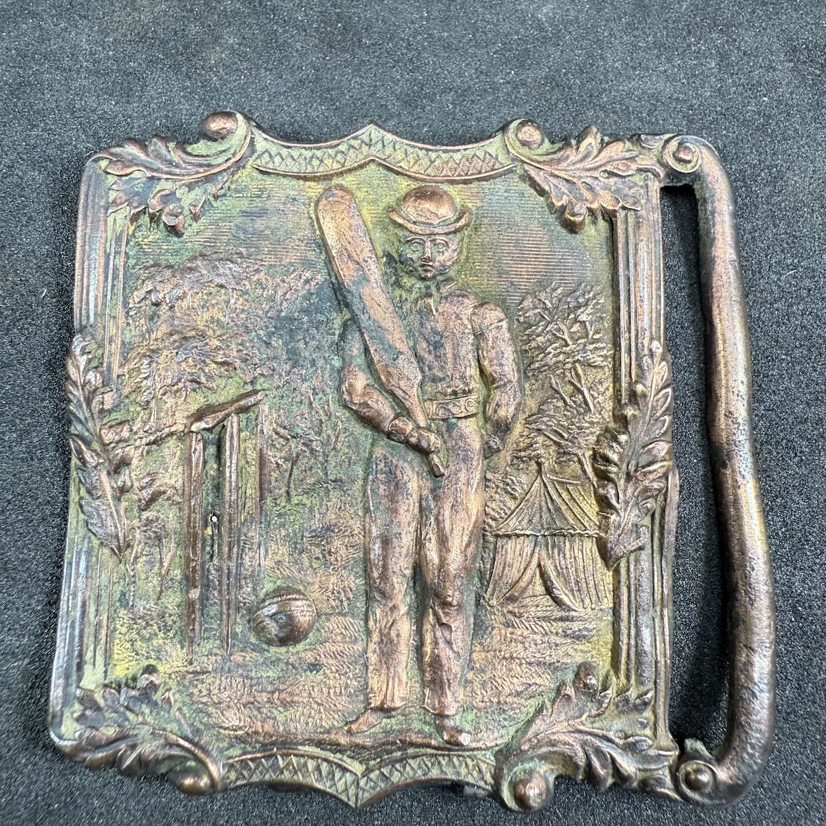 Cricket Belt Sash Buckle. Man in hat holding a bat, large set of stumps with a large ball on the ground, tent and detailed trees as well.