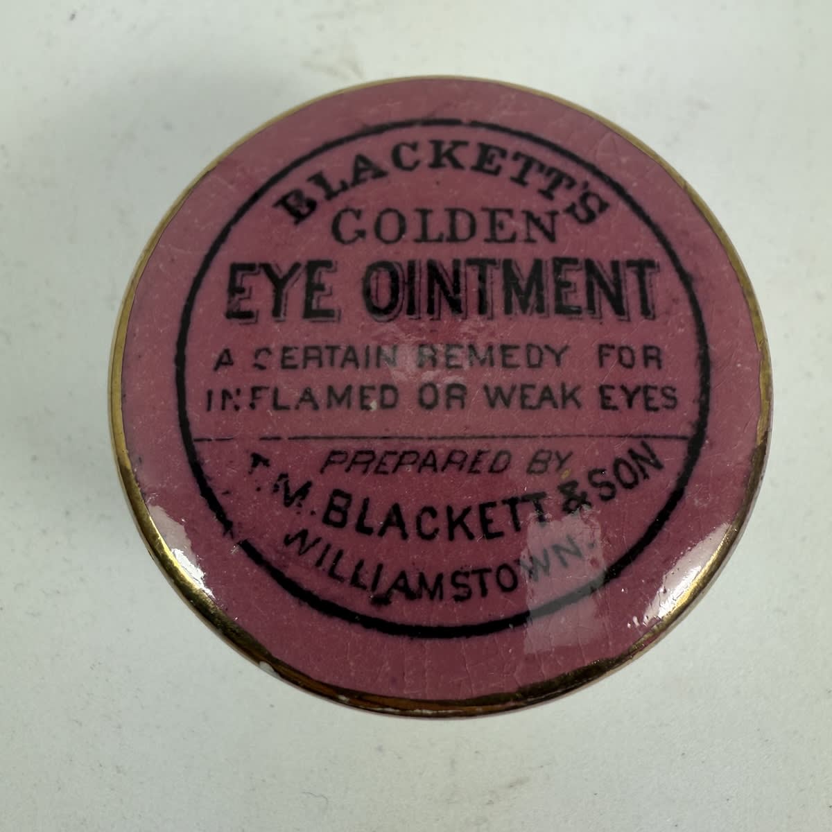 Pot Lid. Blackett's Eye Ointment, Williamstown. T. M. Blackett & Son. Black on Pink. Gold Border version. With fitting base. (Victoria)