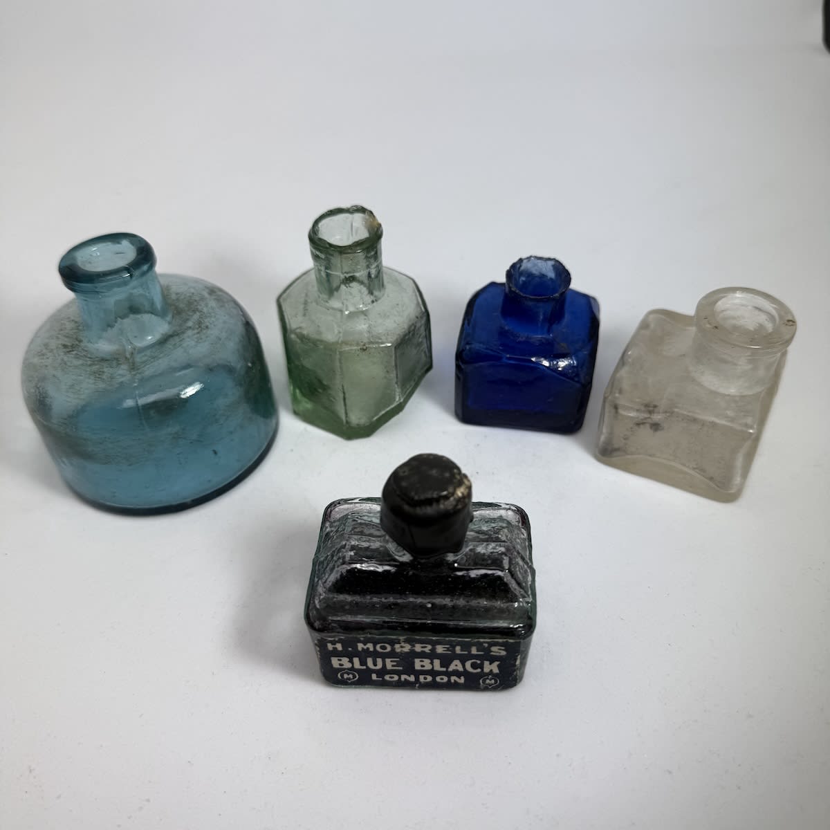 5 Inks: Large round ice blue dome shape; Tipper ink, Octagonal with slanted base; Blue square, facetted shoulder; Clear with pen rest; Boat ink with label Morell's Blue Black London, with some contents.