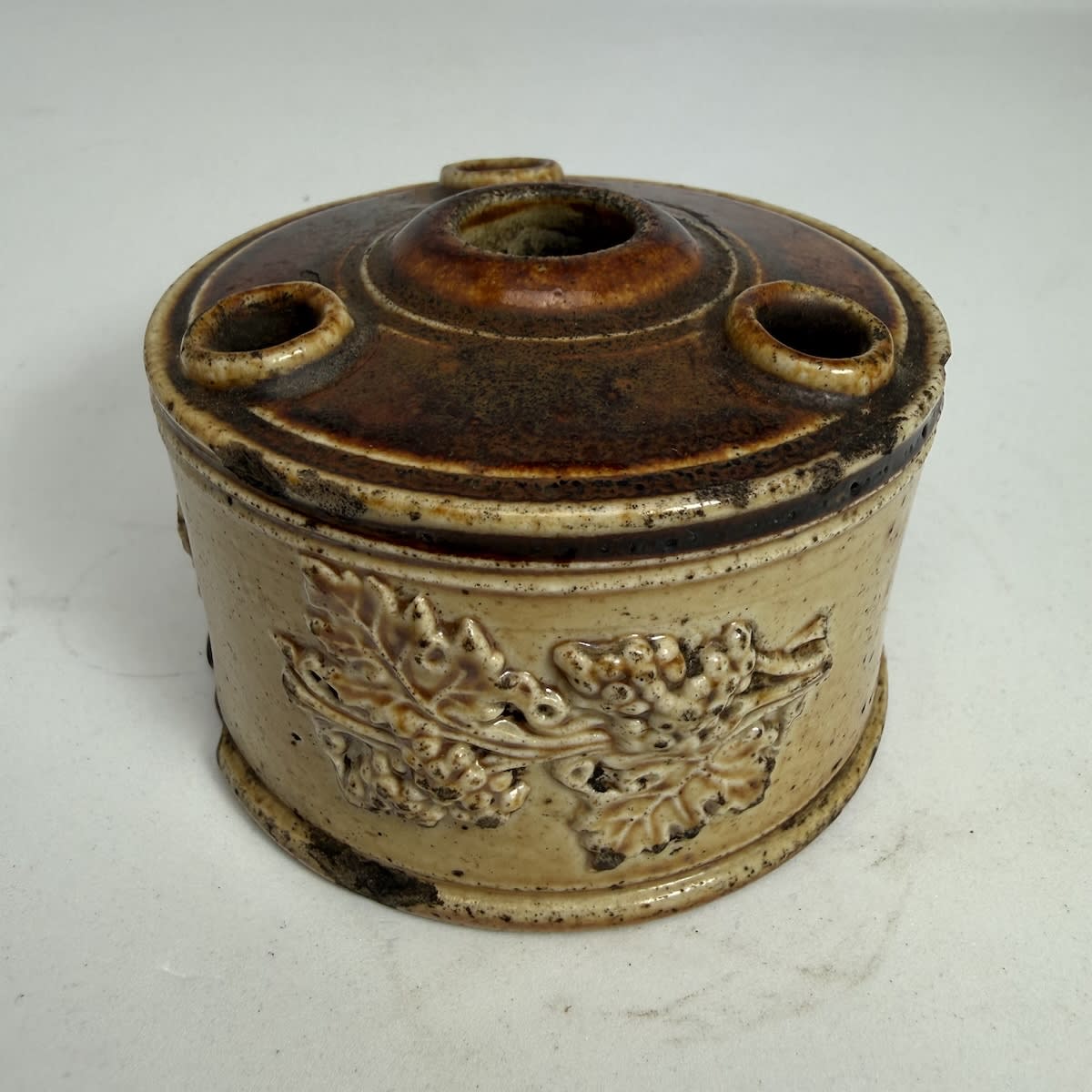 Ink. Round stoneware ink. Dark tan top. Light tan body. Applied Grapes & vine leaf decoration 3 times. no potters stamp.