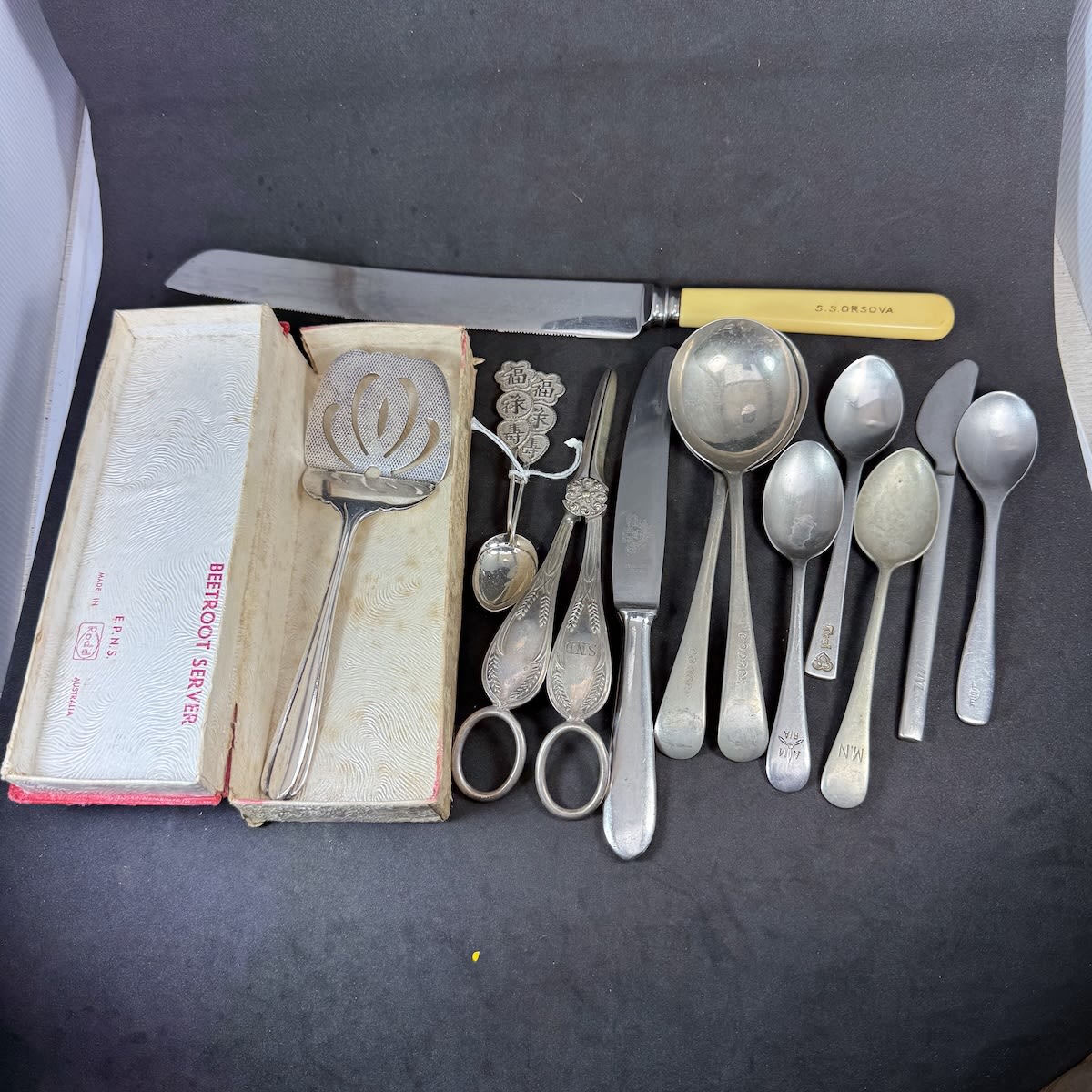 13 Cutlery Pieces: Bread Knife SS Orsova; SND Crimper; Various marked Teaspoons and Knives; 2 teaspoons with chinese symbols; 2 CO DOCK Soup Spoons; Beetroot Server in box.