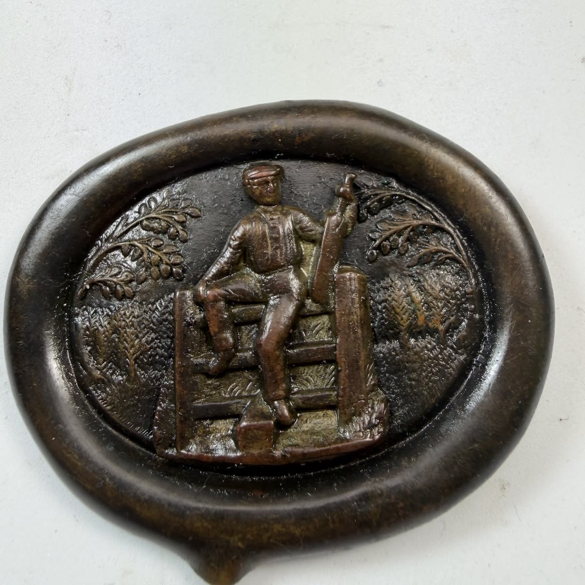Cricket Belt Buckle / Sash Buckle. Oval backing piece with trees, rural setting. Pinned middle piece with a cricketer holding a bat climbing over a gate.