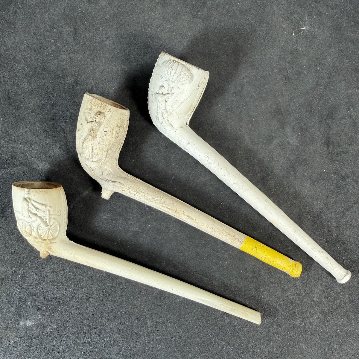 3 Complete Clay Pipes: Man Holding A Parachute; Boxers on either side Sullivan (John L. Sullivan) and ? possibly Mitchell but the name is very poorly struck and could just be Sullivan again; Man on bicycle on two sides.